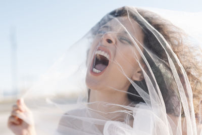 Young angry female with curly hair screaming through transparent veil on sunny clear day