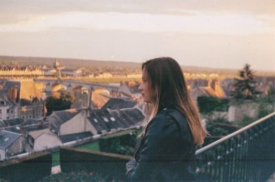 Side view of young woman looking at cityscape while standing on bridge against sky