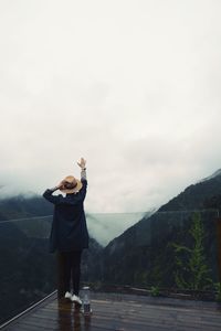 Full length of man with hand raised standing by glass railing against mountains