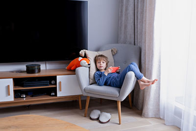 Cute preschool blonde kid using smartphone sitting on a cozy chair in light nordic style living room