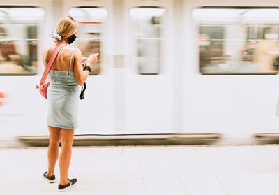 Back view of unrecognizable young female passenger in casual outfit and black protective mask standing on platform of subway station and checking information on mobile phone