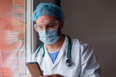 Male doctor using smart phone while standing by window