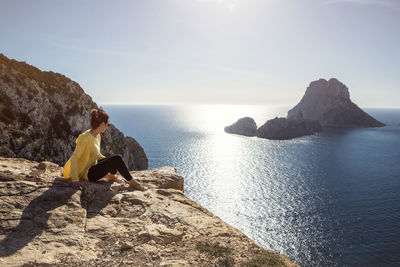 Woman in yellow zip jacket sitting on a cliff looking out to es vedra near ibiza