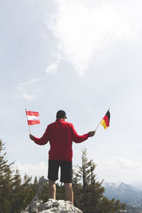 Rear view of mature man holding national flags while standing on mountain against sky