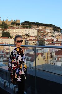 Portrait of smiling young woman standing by railing against cityscape