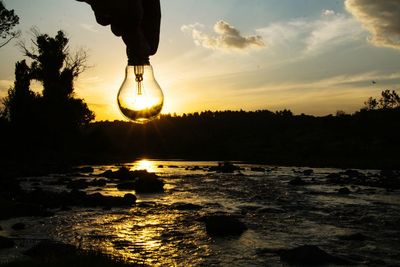 Cropped silhouette hand holding light bulb against river at sunset