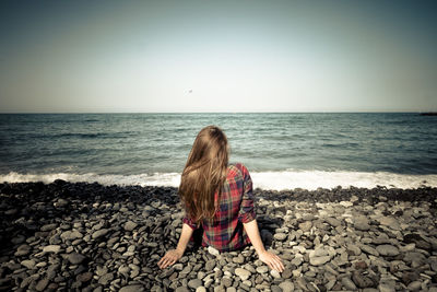 Rear view of woman sitting on rock at beach against clear sky