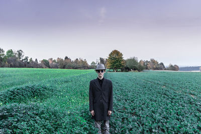 Portrait of young man wearing coat and hat while standing on agricultural field against sky