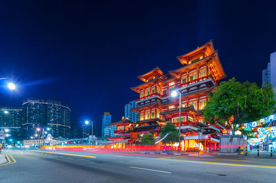 Illuminated road by buildings against sky at night