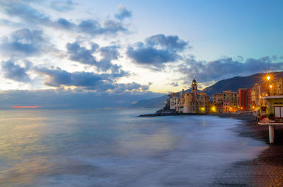 Illuminated buildings by sea against sky during sunset