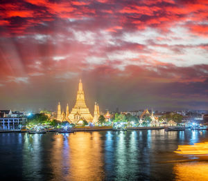 Illuminated buildings at waterfront against cloudy sky