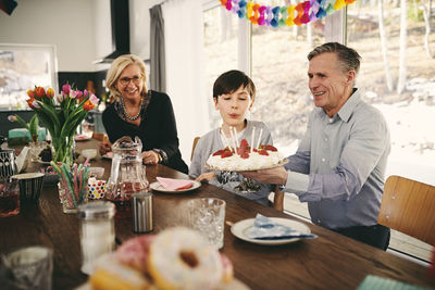 Boy blowing candles on birthday cake while sitting with grandparents at table