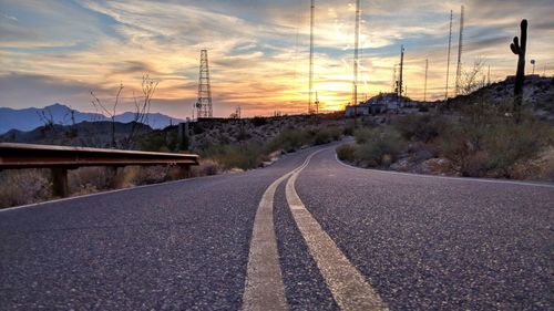 Road by electricity pylon against sky during sunset