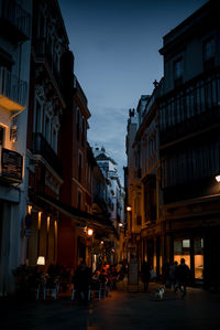 People at sidewalk cafe on street amidst buildings in city at dusk