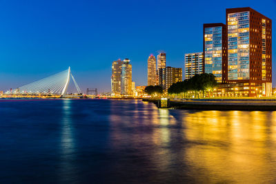 Illuminated erasmus cable-stayed bridge over maas river by buildings against blue sky at night