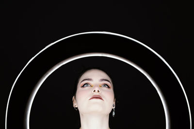 Female with professional makeup and in stylish wear standing in studio behind ring circle lamp on black background and looking away