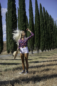 Full length of woman holding hat standing against trees