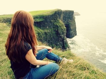 Woman sitting on grass at cliff looking at sea