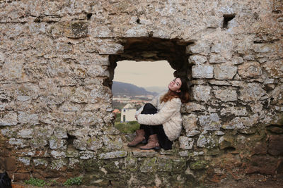 Full length portrait of young woman sitting amidst stone wall at window