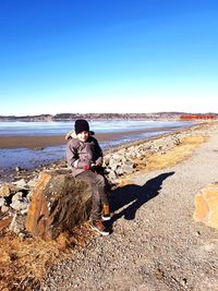 Boy sitting on rock at beach against clear sky