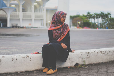 Young woman sitting at roadside against mosque in city