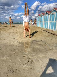 Shirtless man doing handstand on sand at beach against cloudy sky