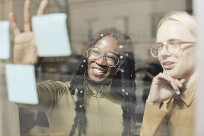 Smiling female entrepreneur explaining strategy to non-binary colleague on adhesive note over glass window in office
