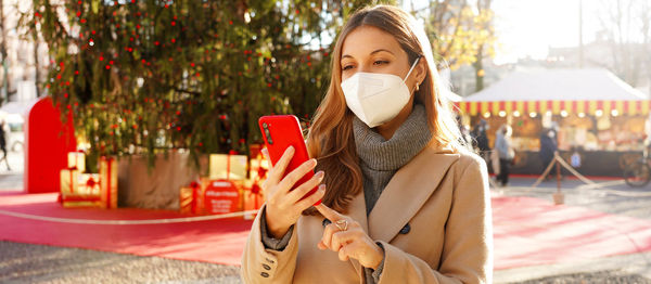 Stylish fashion girl wearing face mask woman typing on smartphone with christmas tree and markets