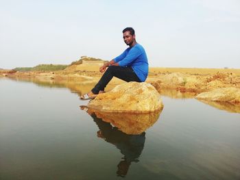 Young man sitting on rock at lake against sky