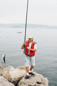 Side view of active man in red waistcoat with fishing rod standing on stony shoe and fishing in bright day