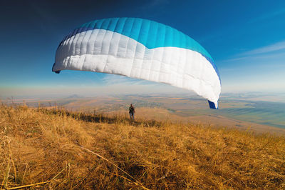 A paraglider takes off from a mountainside with a blue and white canopy and the sun behind. a