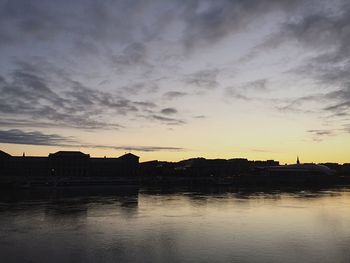 Silhouette buildings by calm lake against cloudy sky