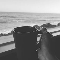 Close-up of hand holding coffee cup by sea against sky