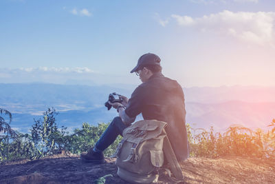 Side view of man photographing while sitting on mountain against sky