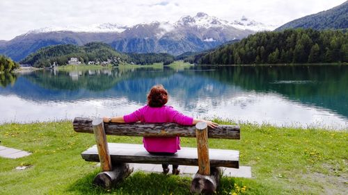 Rear view of woman sitting on bench at lakeshore against mountains