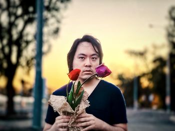 Portrait of young asian man holding red and purple tulips against trees and sunset sky.