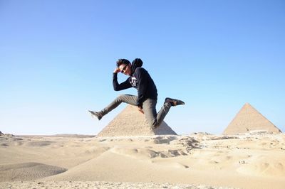 Young man wearing hooded shirt and sunglasses in mid-air at giza pyramids against clear sky
