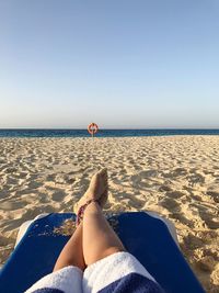 Low section of woman relaxing on lounge chair at sandy beach against clear sky