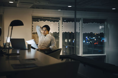 Confident male entrepreneur reading document while sitting in dark office at night