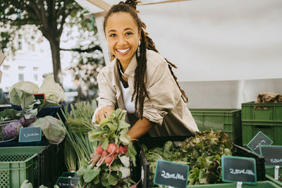 Portrait of happy female owner holding vegetable near crates in stall at market