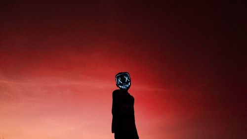 Low angle view of boy standing against sky during sunset with a spooky face mask