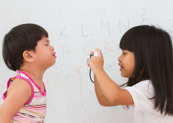 Side view of girl photographing brother crying against wall