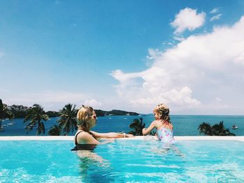 Woman with daughter swimming in pool by sea against cloudy sky