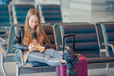 Full length of girl using phone while sitting in waiting room at airport