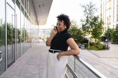 Smiling young businesswoman with hand in pocket talking on mobile phone by railing