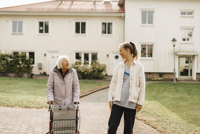 Smiling female nurse and senior woman looking at each other while walking against house