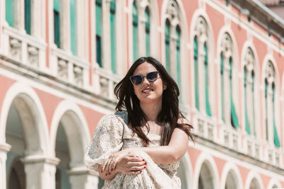 Portrait of beautiful young woman with long dark hair, sitting in square with beautiful architecture