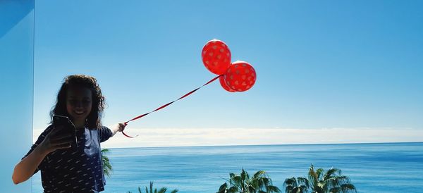 Smiling young woman using mobile phone while holding red balloons against sea