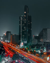 Light trails on street amidst illuminated buildings against sky at night