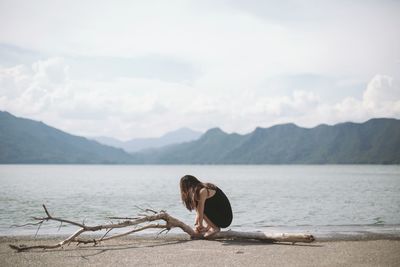 Girl sitting on driftwood log at beach against mountain range
